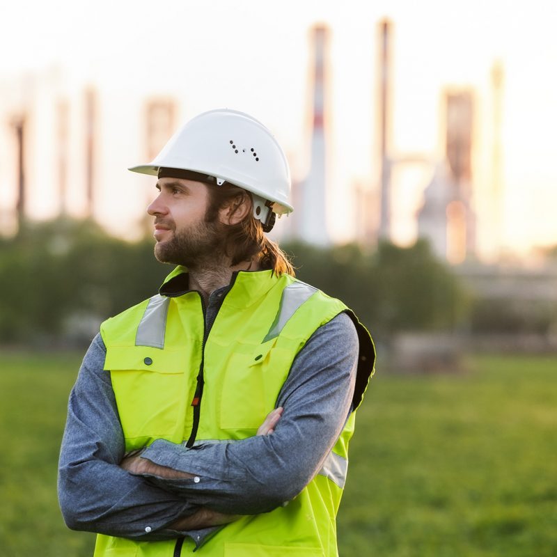 Portrait of young engineer standing outdoors by oil refinery, arms crossed.